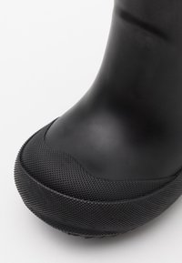 Botte en caoutchouc noir avec une section inférieure texturée, une partie supérieure lisse, une pointe arrondie et un capuchon de protection renforcé. Le matériau est durable et imperméable.