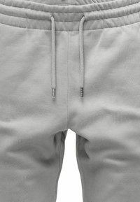 Hellgraue Sweatpants mit elastischem Bund, verstellbarem Kordelzug und glatter Textur. Mit silbernen Metallakzenten an den Kordelzügen.