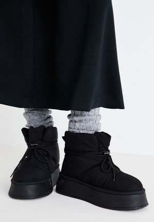 Bottines noires matelassées avec semelles épaisses portées par-dessus des chauffe-jambes en maille grise et une longue jupe noire.