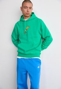 Hanorac verde cu buzunar frontal, prezentând o grafică colorată. Purtați cu pantaloni de trening albaștri, care arată un logo alb Nike.