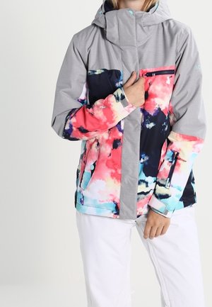 Person trägt eine graue Kapuzenjacke mit pinken, blauen und schwarzen abstrakten Mustern sowie weiße Hose, Hand nahe der Brusttasche mit Reißverschluss.