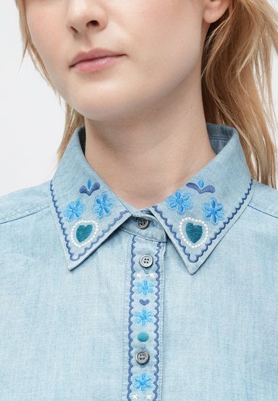 Primo piano di un colletto di camicia blu chiaro con ricami floreali e cuori blu, abbottonato con tre bottoni visibili sulla pattella.