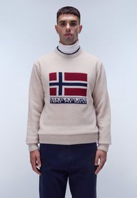 Maglione a maglia beige con un design della bandiera norvegese in rosso, blu e bianco, con polsini e orlo a coste; indossato sopra un dolcevita bianco.