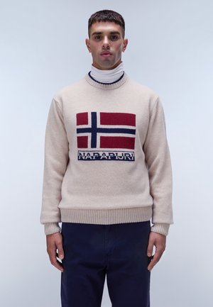 Beige gebreide trui met een Noors vlagontwerp in rood, blauw en wit, met geribde boorden en onderkant; gedragen over een witte coltrui.