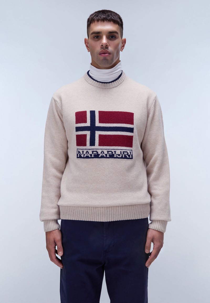 Maglione a maglia beige con un design della bandiera norvegese in rosso, blu e bianco, con polsini e orlo a coste; indossato sopra un dolcevita bianco.