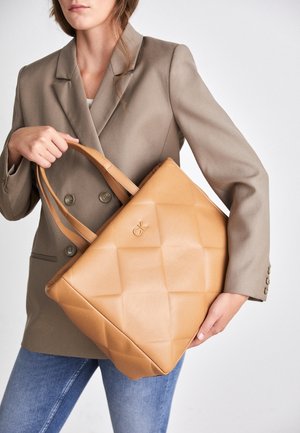 Vrouw in beige blazer en spijkerbroek met een tan gekwartste leren totebag met het CK-logo aan de voorkant, tegen een witte achtergrond.