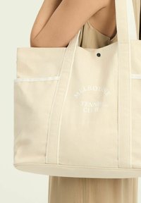 Beżowa torba canvas z białymi akcentami, z napisem "Melbourne Tennis Club" wydrukowanym z przodu i z zapięciem na guzik u góry.