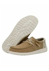 Zapatilla de lona marrón con suela de goma blanca. Presenta un panel de talón texturizado en contraste y un diseño de cordones con una etiqueta decorativa.