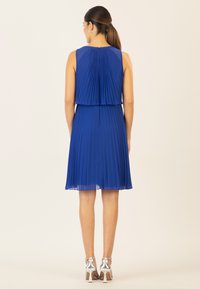 Blaues plissiertes Kleid mit tailliertem Oberteil, geschichtetem Design und ausgestellt Rock. Silberne High Heels ergänzen das Outfit.