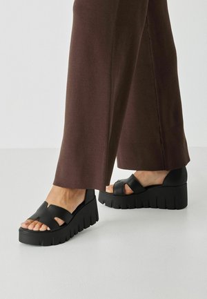 Bare fødder iført sorte platformsandaler med brede remme, parret med brune bukser med vide ben mod en ensfarvet baggrund.