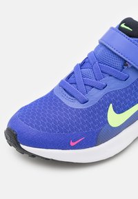 Zapato deportivo azul con parte superior de malla, cordones azules y logotipos de swoosh de Nike en verde neón y rosa sobre suela blanca y punta negra.