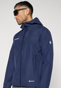 Mörkblå vattentät jacka gjord av GORE-TEX-material, med huva, fickor med dragkedja och en slank design med logotypdetaljer.