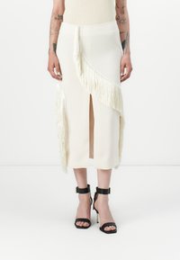 Jupe midi crème avec un détail en franges sur un côté et une fente à l'avant, associée à des sandales noires à talons avec des brides autour de la cheville.