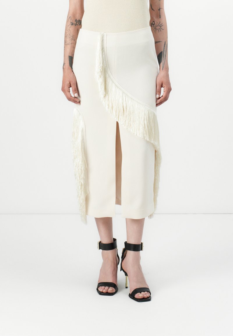 Jupe midi crème avec un détail en franges sur un côté et une fente à l'avant, associée à des sandales noires à talons avec des brides autour de la cheville.