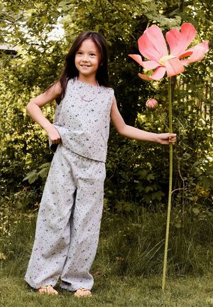 Smilende jente i løs, blomster-mønstret antrekk holder en høy rosa blomst med store kronblader foran en grønn hagebakgrunn.