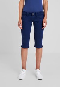 Žena v námořnických modrých zkrácených kalhotách a bílých teniskách stojí na pozadí jednoduchého světlého šedého pozadí.