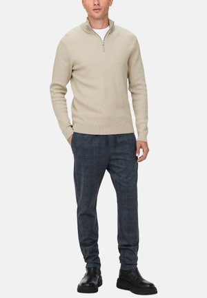 Beige gebreide pullover met een opstaande kraag en een halve rits, gecombineerd met donker geruite broeken en zwarte enkelboots.