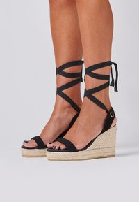 Zwarte espadrille wedges met kruislings enkelbandjes, een van jute gewikkelde hak en een open teen. Beschikt over een gladde bovenkant en een gestructureerde zool.