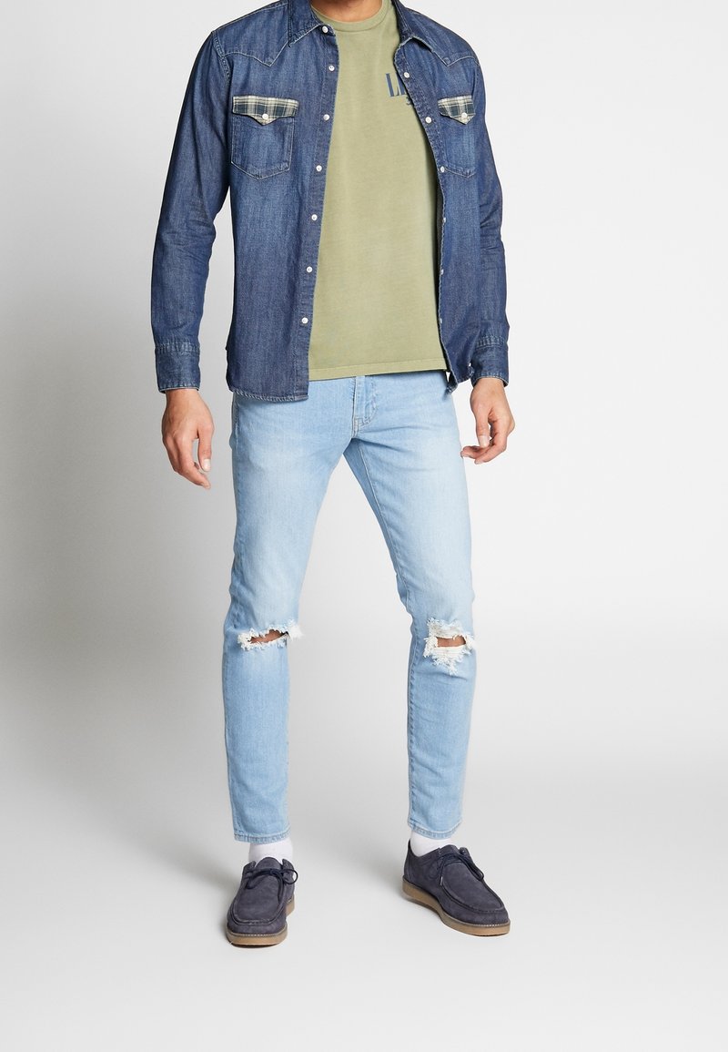 Homme portant une chemise en jean bleue sur un t-shirt vert olive, un jean clair déchiré, des chaussettes blanches et des chaussures en daim bleu foncé, debout sur un sol gris.