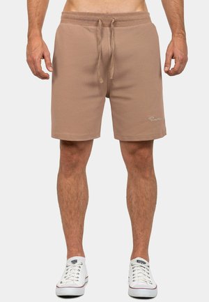 Hombre vestido con pantalones cortos marrones con cordón hasta la rodilla y zapatillas blancas bajas, de pie contra un fondo liso.