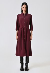 Robe bordeaux avec col, manches longues, fermeture à boutons et une ceinture assortie ; fabriquée en tissu lisse. Portée avec des bottes à talons noires.