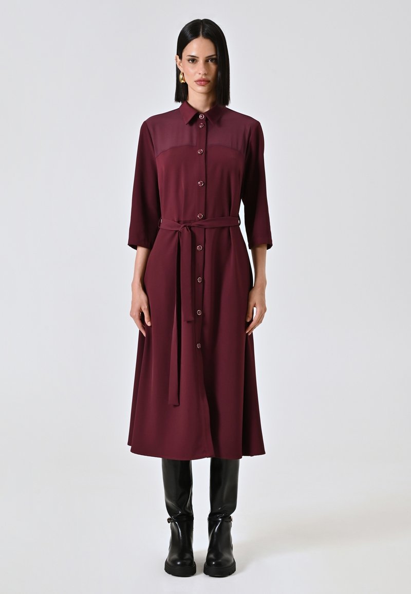 Robe bordeaux avec col, manches longues, fermeture à boutons et une ceinture assortie ; fabriquée en tissu lisse. Portée avec des bottes à talons noires.