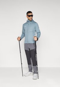 Lys blå zip-up jakke med hætte, grå og sorte bukser samt vandrestave. Den har tekstureret stof og en praktisk design til udendørs brug.