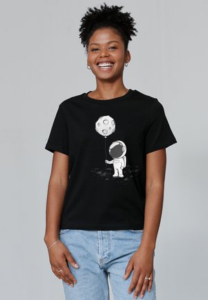 Schwarzes Baumwoll-T-Shirt mit einem weißen Grafikmotiv eines Astronauten, der einen Mondluftballon hält. Das Model trägt hellblaue Jeans und hat kurze Ärmel.