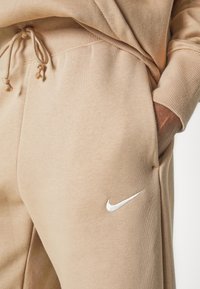 Beige bomullsträningsbyxor med snörmidja, sidfickor och en liten vit Nike-logotyp på vänster lår. Slät textur.