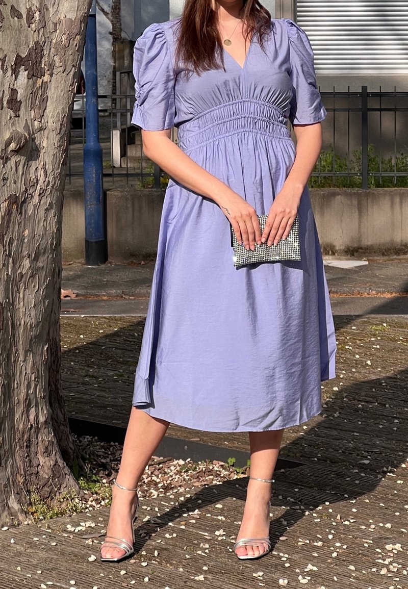 Femme debout à l'extérieur portant une robe midi violet clair, tenant une petite pochette argentée et portant des talons ouverts argentés.