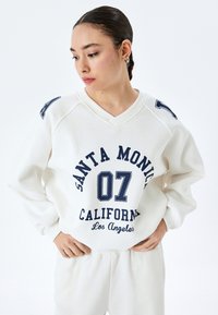 Felpa bianca con scollo a V, caratterizzata da una stampa grafica blu con la scritta "SANTA MONICA 07 CALIFORNIA LOS ANGELES." Tessuto morbido, vestibilità comoda.