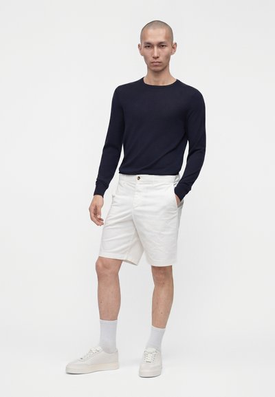 Homme debout portant une chemise noire à manches longues, un short blanc aux genoux, des chaussettes blanches et des baskets blanches sur un fond uni.