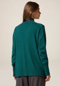 Maglione verde con orlo e polsini a coste, caratterizzato da una texture liscia e una vestibilità ampia. Il retro presenta un design semplice senza ulteriori dettagli.