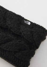 Bonnet en tricot noir avec un épais motif en câble, arborant une petite étiquette de logo intitulée "THE NORTH FACE" sur le côté.