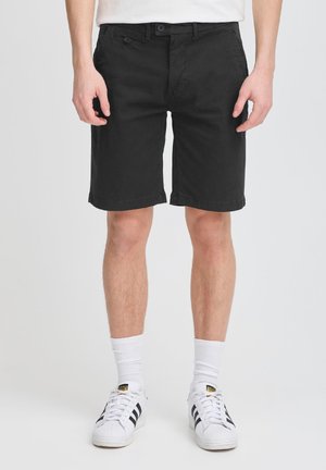 Bărbat purtând pantaloni scurți negri până la genunchi, șosete albe și adidași albi Adidas cu dungi negre pe fundal alb.