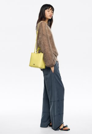 Suéter marrón peludo, pantalones anchos azul marino y bolso de cuero amarillo. Las sandalias tienen correas multicolores. Diseño minimalista, ajuste relajado.