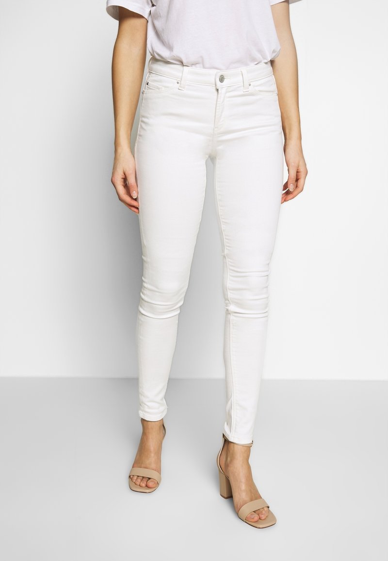Jeans blancos ajustados de cintura alta, con lazos para el cinturón, bolsillos frontales y una textura suave. Combinados con sandalias de tacón bloque en color nude.