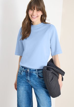Une femme souriante portant une chemise à manches courtes bleu clair et un jean bleu, tenant une doudoune noire sur un fond neutre.