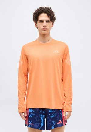 Jeune homme debout, portant une chemise à manches longues orange et un short de sport bleu à motifs, avec un fond clair uni.