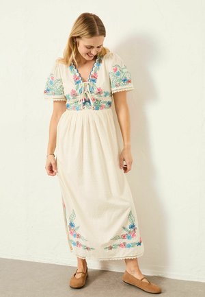Woman wearing a white midi dress with colorful floral embroidery, short sleeves, and lace-up neckline, paired with brown flat shoes, standing indoors.