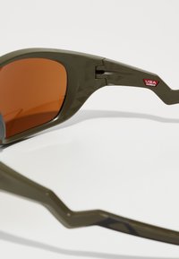 Grüne Sonnenbrille mit eckiger Form, orangen Gläsern, strukturiertem Rahmen und rotem "USA Assembled"-Schild an der Seite.