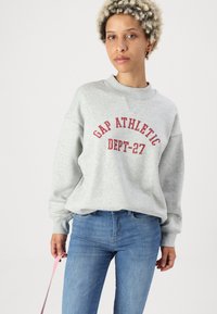 Grå hettegenser med rød "GAP ATHLETIC DEPT-27" tekst, har senkede skuldre, kombinert med lyseblå jeans og en rosa stropp i hånden.
