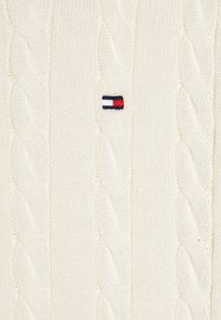 Maglione crema a maglia cable con un piccolo logo ricamato in rosso, bianco e blu. Il tessuto ha un aspetto morbido e testurizzato.