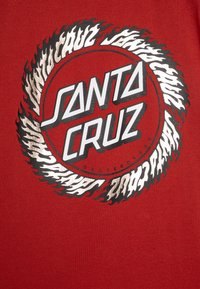 Sarkans T-krekls ar apļa grafiku, kurā melnā un baltā krāsā ir uzraksts "SANTA CRUZ", ko ieskauj liesmām līdzīgas akcenti baltā un melnā krāsā.