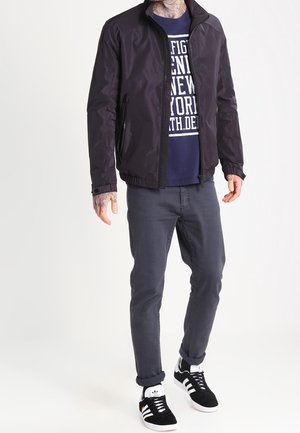 Mężczyzna w czarnej bluzie na zamek, granatowej koszulce z nadrukiem, ciemnych podwiniętych dżinsach i czarnych butach Adidas z białymi paskami.