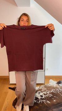 Egy nő maroon színű Anna Field pólót tart az előtte, egy fából készült padlón áll, közel egy tehénbőr szőnyeghez, egy világos szobában.