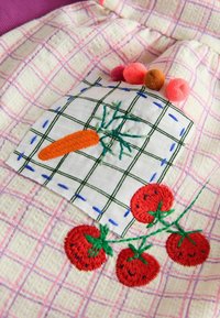 Parche de tela blanca con una zanahoria naranja bordada y tomates rojos sonrientes sobre tela rosa y blanca a cuadros, decorado con cuatro pompones de colores.