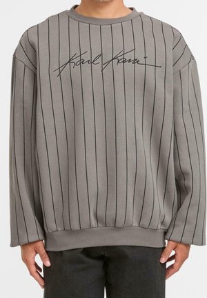 Harmaa collegepaita, jossa on ohuita mustia pystysuoria raitoja ja mustaa kaunokirjoitusta, jossa lukee "Karl Kani" rinnassa. Paita on päällä suorassa seisovalla henkilöllä.