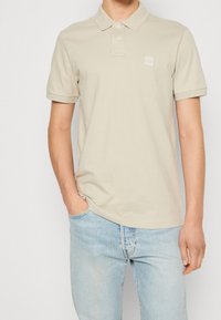Polo beige à manches courtes, avec un col et une petite patch logo. Associé à un jean bleu clair. Design minimaliste.