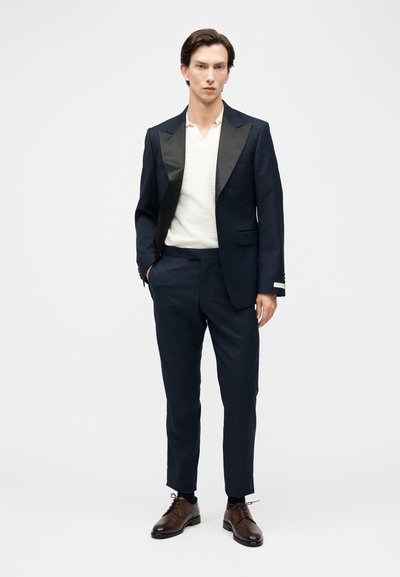 Bărbat purtând un smoking bleumarin cu rever negru, cămașă albă, pantaloni bleumarin și pantofi eleganți maro din piele, stând cu o mână în buzunar.